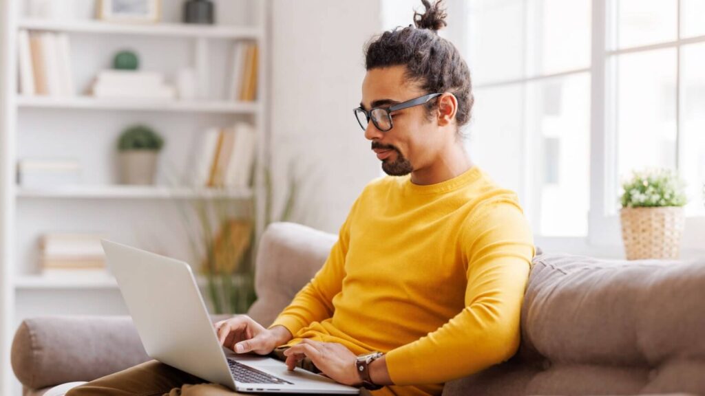 A man in a yellow sweater sitting on a couch and working on a laptop.