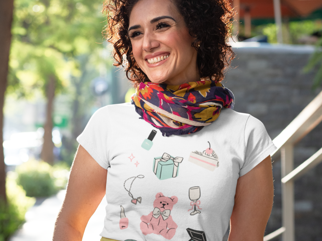 Smiling woman outdoors, wearing a white t-shirt with colorful doodles of makeup, a gift box, cake, and a scarf.