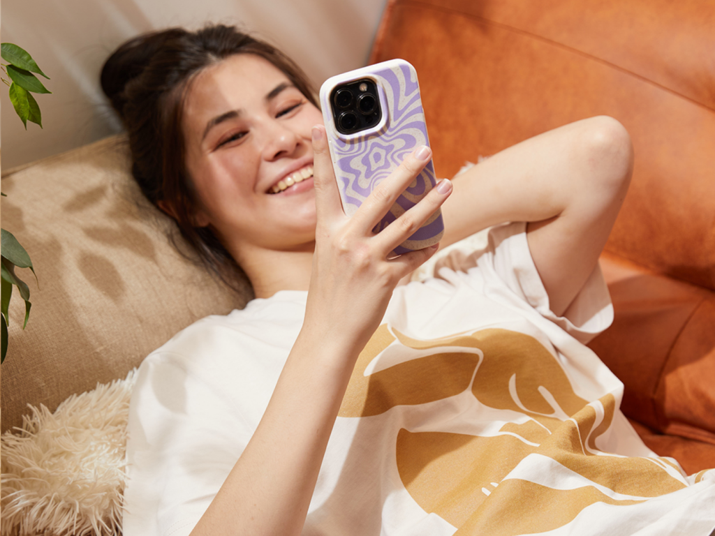 A woman lies on a couch, smiling at her phone with a purple-patterned case. She's wearing a cream shirt with a gold print.