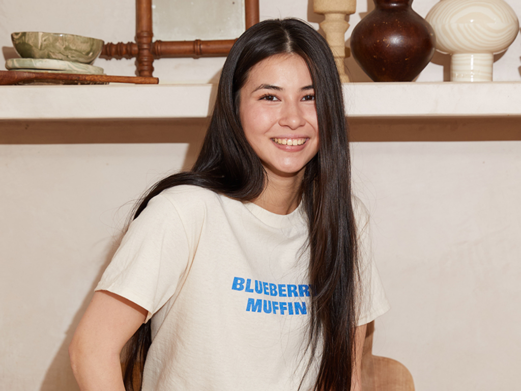 A woman with long dark hair is smiling, wearing a cream "Blueberry Muffin" t-shirt. Behind are decorative ceramics on a wooden shelf.