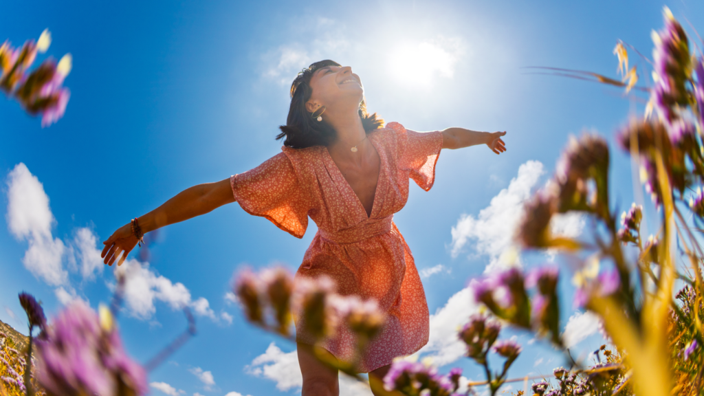 2 6 Custom summer apparel for seasonal sales A joyful woman in a red floral dress spreads her arms under a bright sunlit sky, surrounded by purple flowers and a few clouds, conveying freedom and happiness.