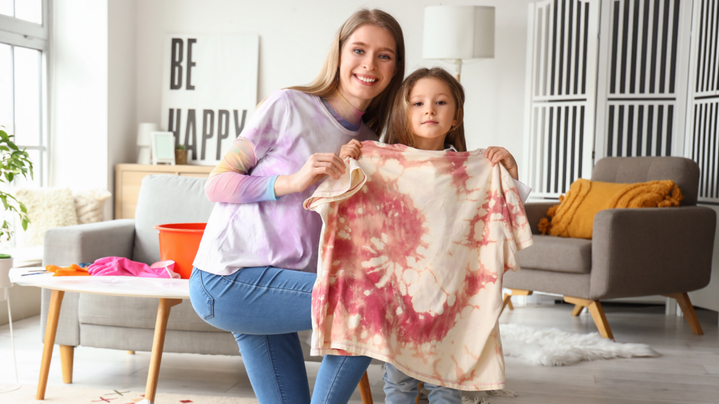 4 Simple tie dye patterns to jumpstart your collection A woman and a child happily display a tie-dyed shirt in a cozy, bright living room with modern decor, promoting a joyful and creative atmosphere.