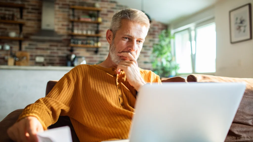 5 Tips to successfully sell shirts on Etsy A middle-aged man in an orange sweater sits on a couch, thoughtfully looking at a laptop in a cozy, sunlit living room.