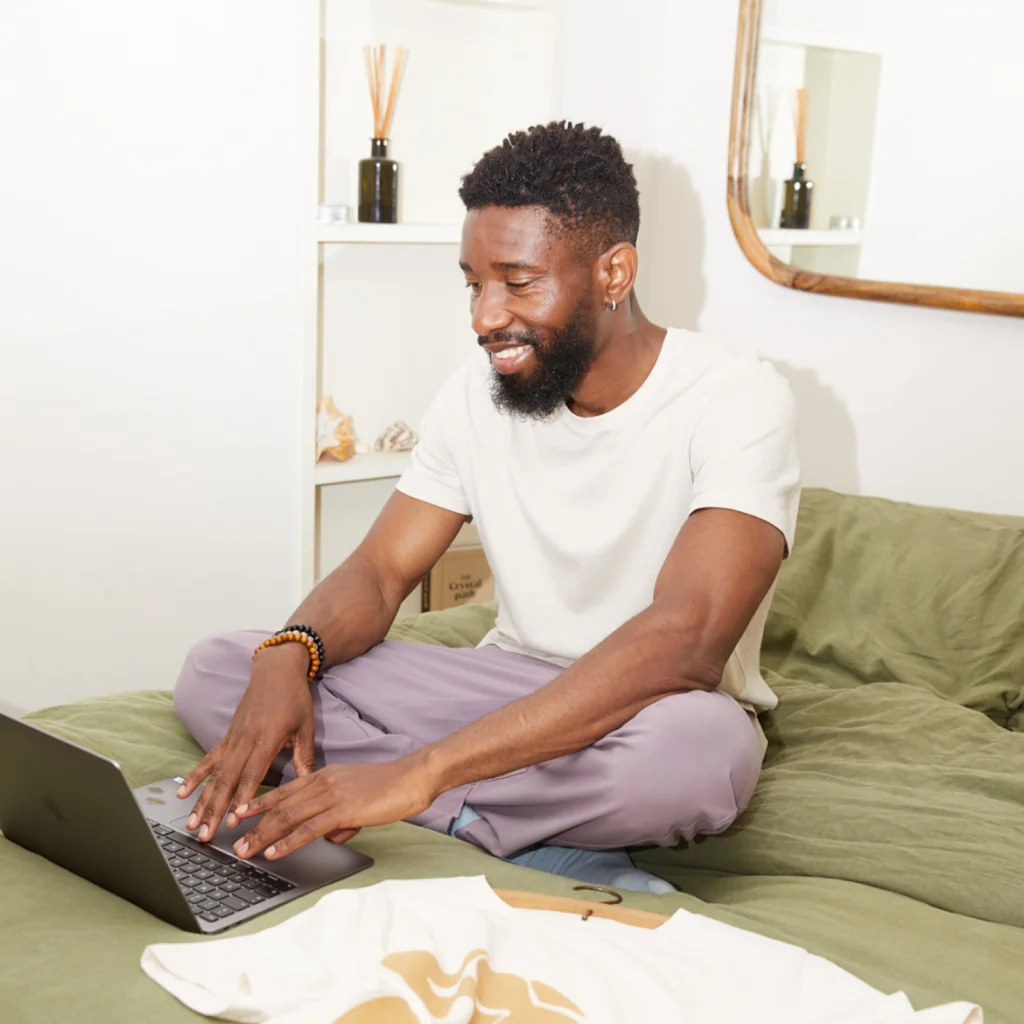 Define your target audience 1 A man with a beard smiles while using a laptop to learn how to sell t-shirts on Etsy, sitting cross-legged on a bed.
