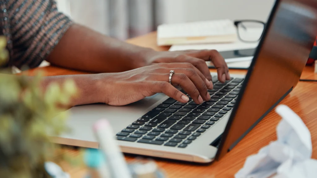 Hands typing on a laptop keyboard on a wooden desk, surrounded by office items like papers and a plant.