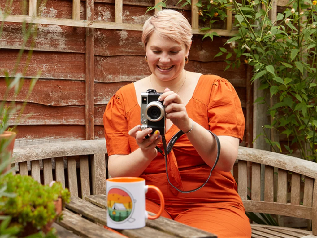 A smiling woman in an orange dress takes a picture of a colorful mug on the table, with greenery surrounding her.