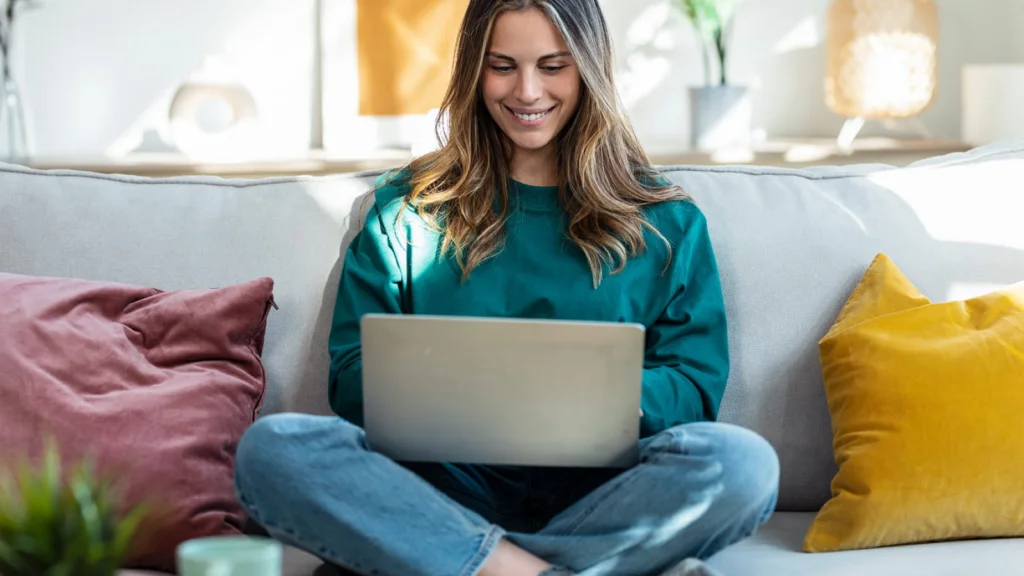 A smiling woman in a green sweater sits cross-legged on a sofa, using a laptop to search for Etsy shop name ideas.