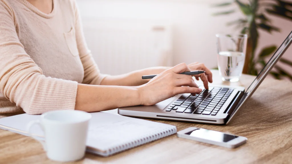 Set up your Etsy storefront A person types on a laptop at a wooden desk, surrounded by a notebook, coffee mug, smartphone, and glass of water.