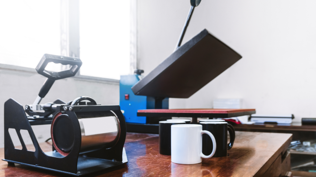 Heat press machine with open platen on a wooden table, next to a mug press holding a white mug.